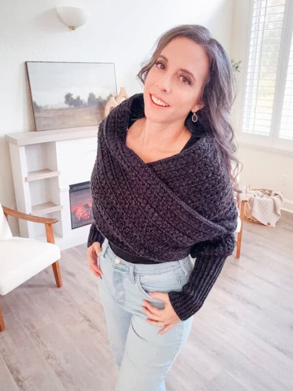 Person with long dark hair wearing a textured black wrap sweater and light jeans stands in a room with a fireplace and chair.