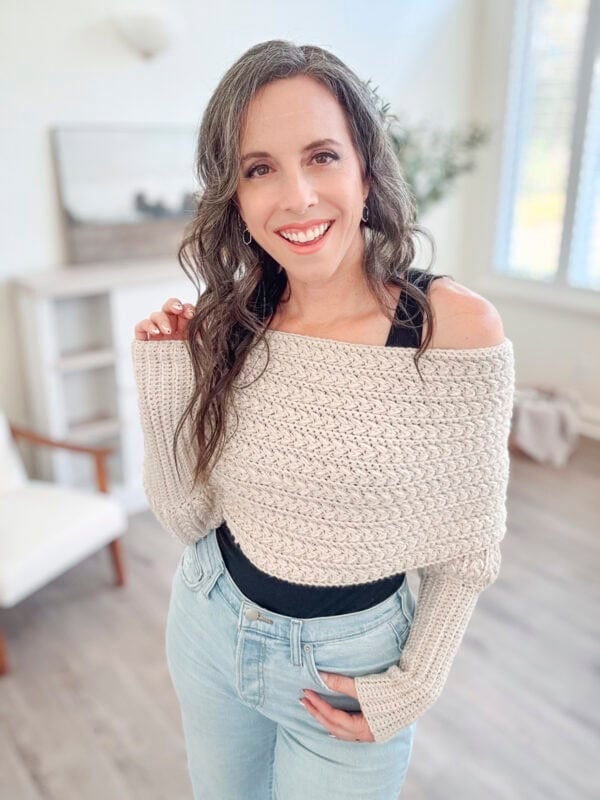 A woman with long, wavy hair in a beige knit sweater and jeans stands smiling in a bright room with a soft, neutral color scheme.