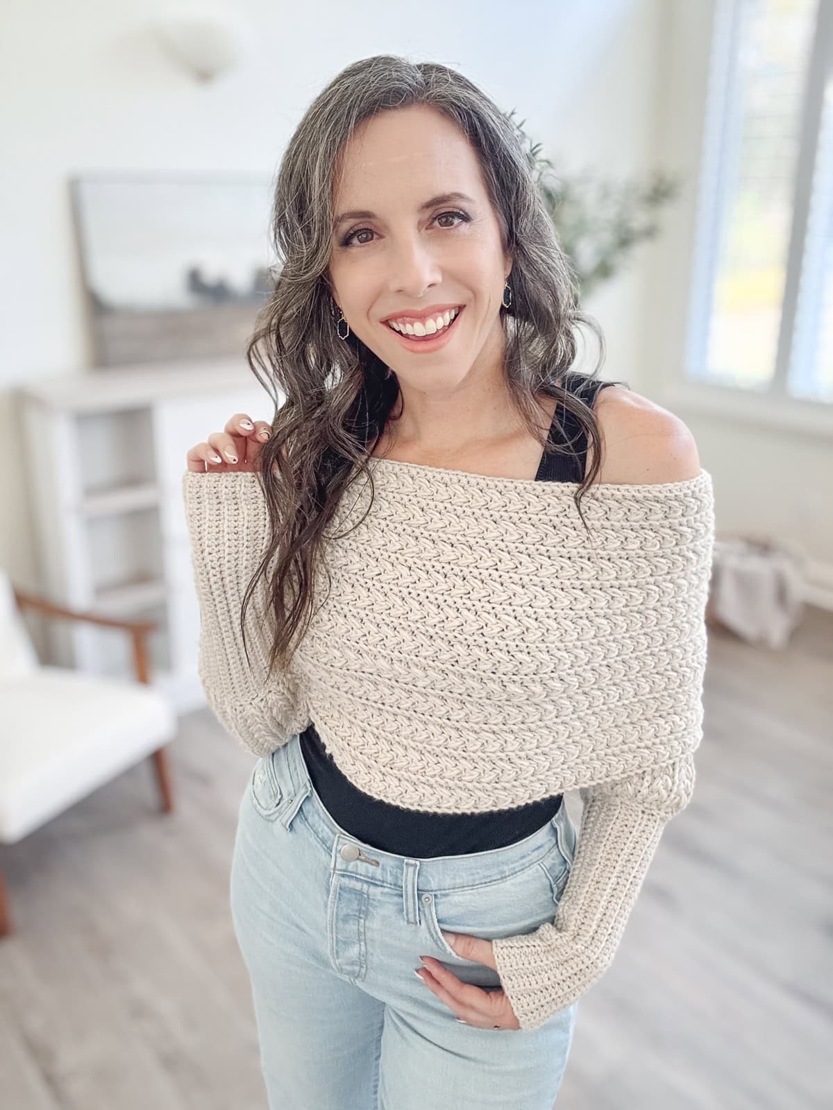 A woman with long, wavy hair in a beige knit sweater and jeans stands smiling in a bright room with a soft, neutral color scheme.