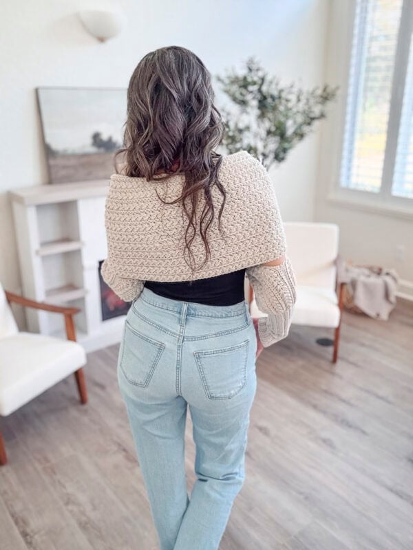 A woman with long hair faces away, wearing a beige knit sweater and light blue jeans. She stands in a modern, bright room with chairs and a plant.