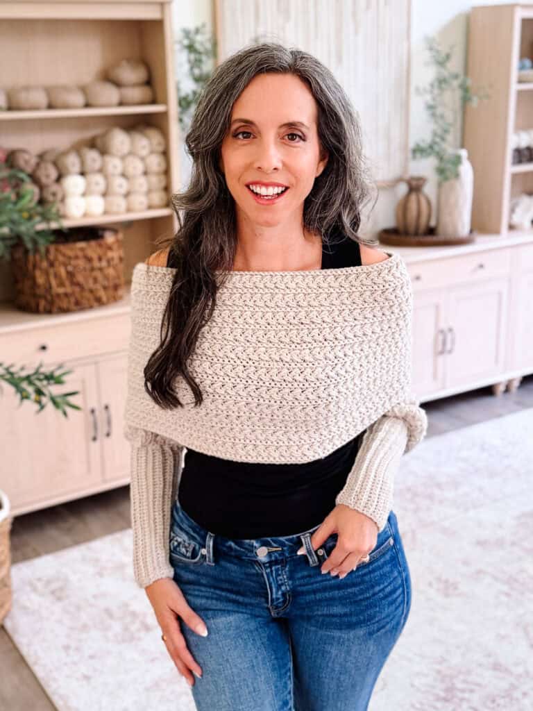 Woman with long gray hair wearing a cream knit shawl and jeans stands indoors, smiling at the camera. Shelves with yarn and decor are visible in the background, giving the cozy space an inviting feel—perfect for showcasing her Winter Wheat Crochet Sweater Scarf Pattern.