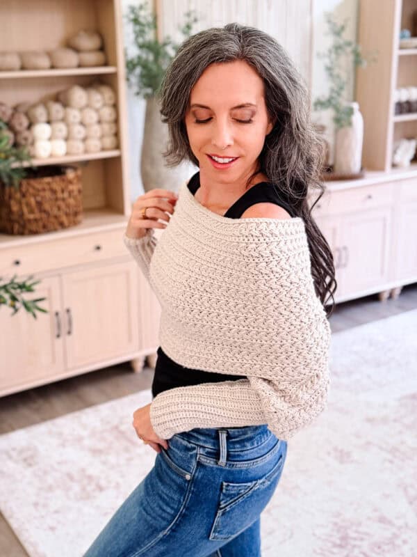 Woman with long gray hair wears a cream crocheted sweater, inspired by the Winter Wheat Crochet Sweater Scarf Pattern, and jeans, standing in a cozy, well-lit room with shelves and yarn in the background.