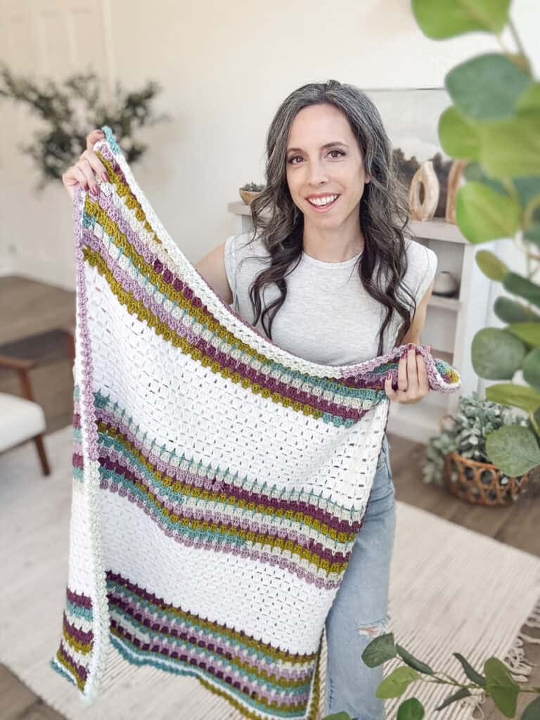 A person holds a colorful, striped crochet blanket in a room with plants and neutral decor.