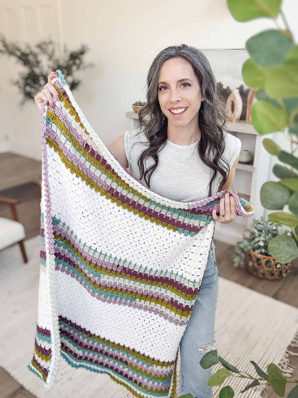 A person holds a colorful, striped crochet blanket in a room with plants and neutral decor.