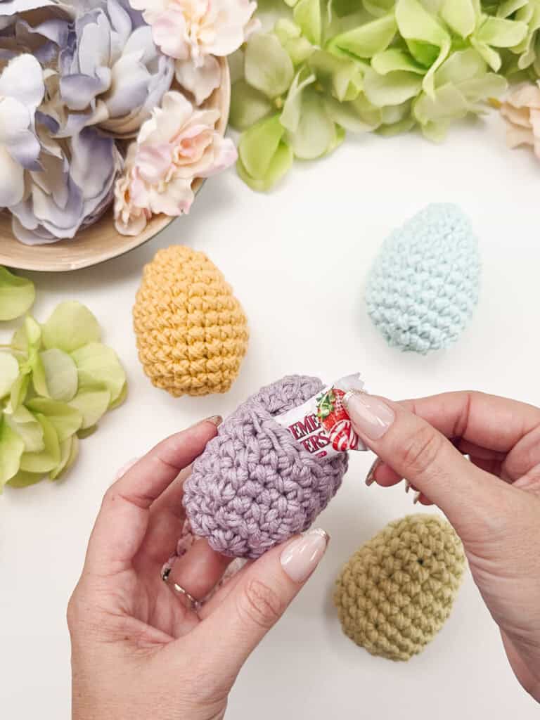 Hands placing candy in a purple crocheted egg, surrounded by green, yellow, light blue, and light green crocheted eggs, with flowers in the background.