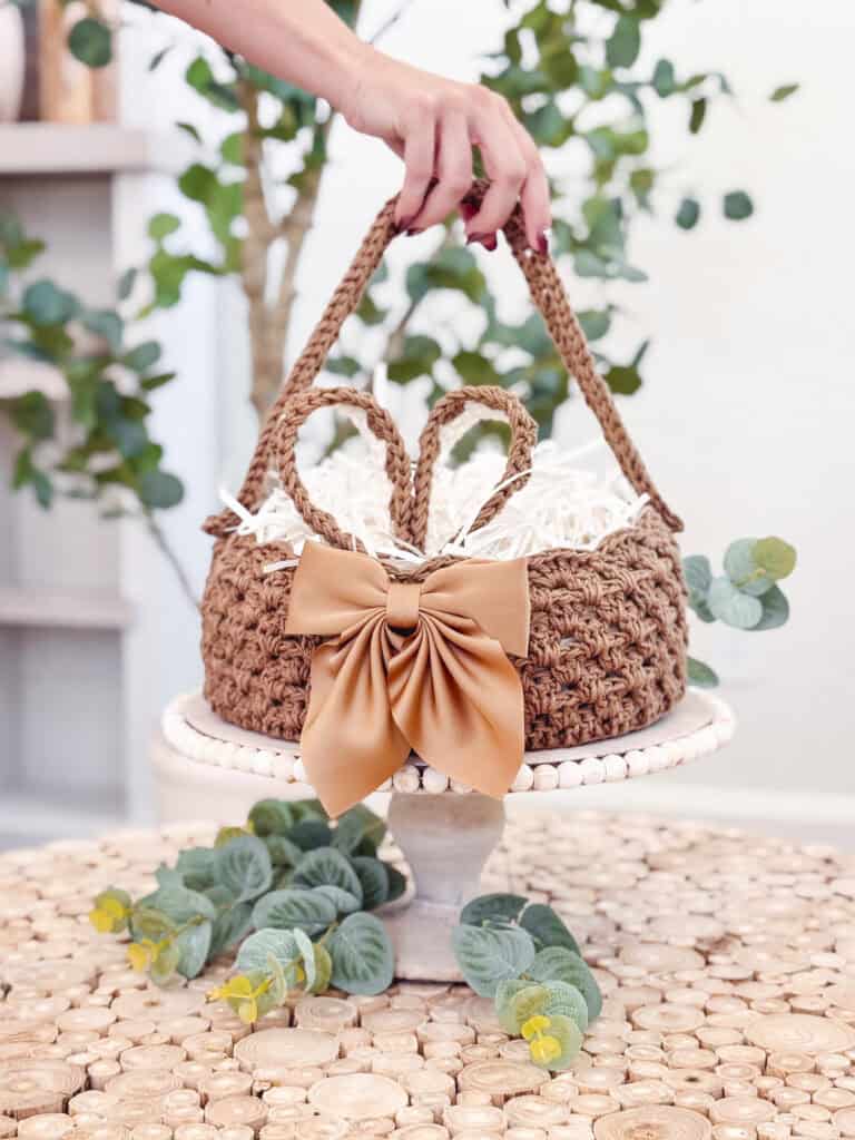 A hand holds a woven basket with a large bow, filled with shredded paper. The basket sits on a pedestal, surrounded by green foliage, on a wooden surface.