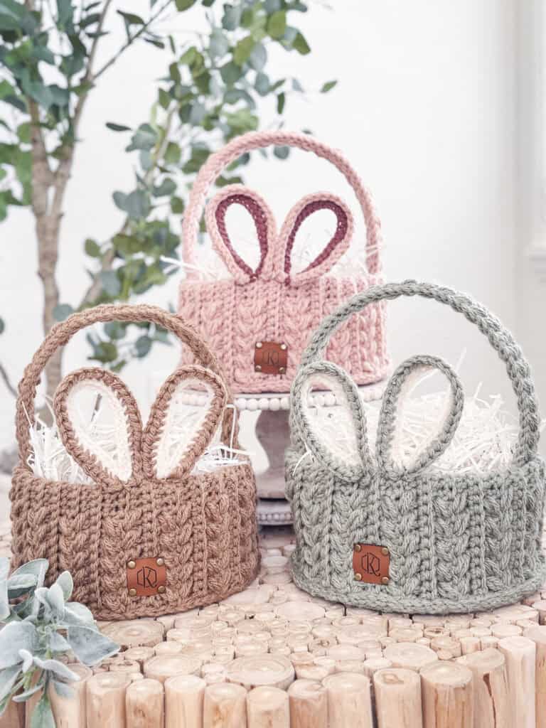 Three knitted baskets with bunny ear designs in pink, brown, and gray shades, placed on a wooden surface with greenery in the background.