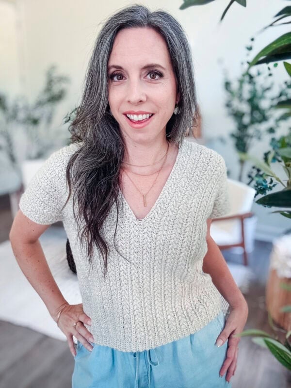 A woman with long gray hair wearing a Braided Wheat Easy Summer Crochet Top Pattern and light blue pants stands indoors, smiling at the camera. Plants and furniture are visible in the background.