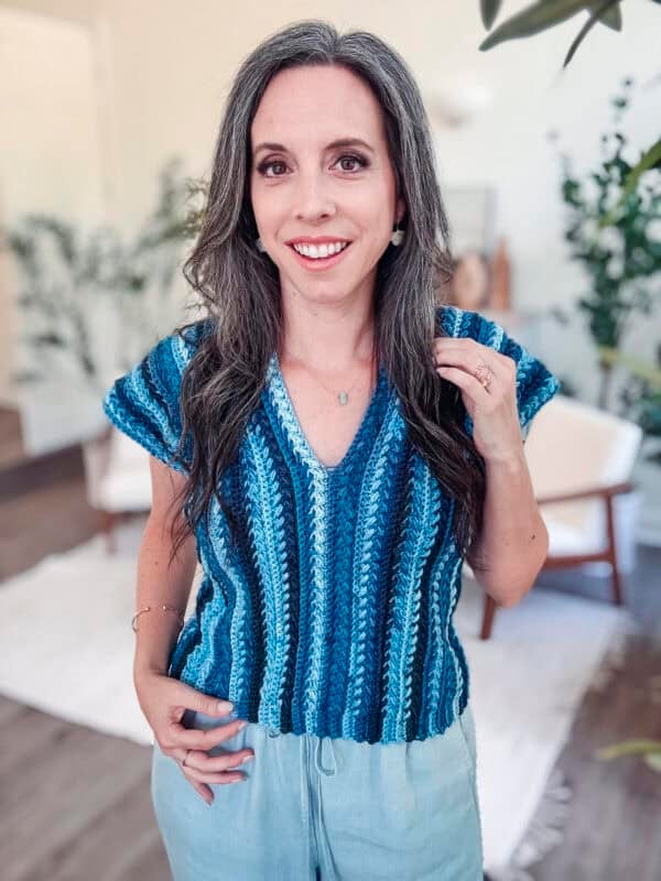 A woman with long gray hair wearing a blue and white striped knit top, similar to the Braided Wheat Easy Summer Crochet Top Pattern, and light pants stands indoors, smiling at the camera.