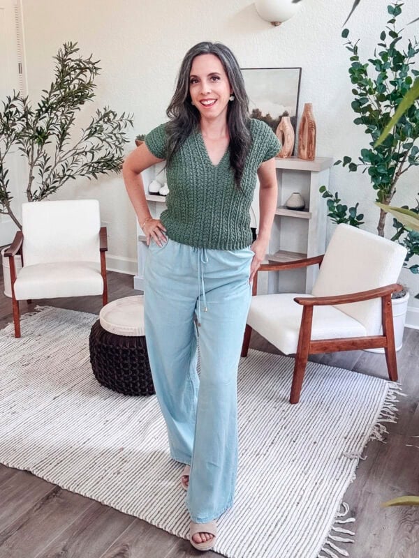 Person with long hair smiles, standing in a room with white chairs, a rug, and plants. They wear a green top and light blue pants.