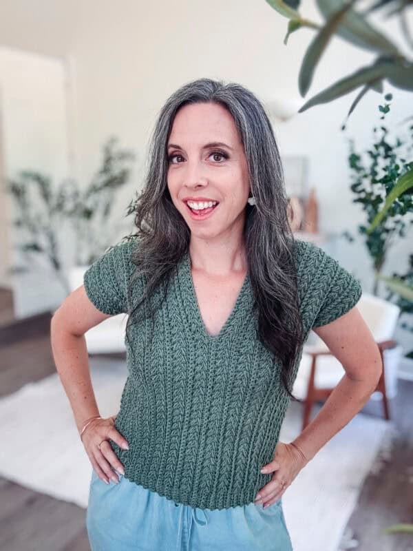 A woman with long, gray hair and a green Braided Wheat Easy Summer Crochet Top Pattern stands indoors, smiling at the camera with her hands on her hips. The background shows plants and light-colored furniture.