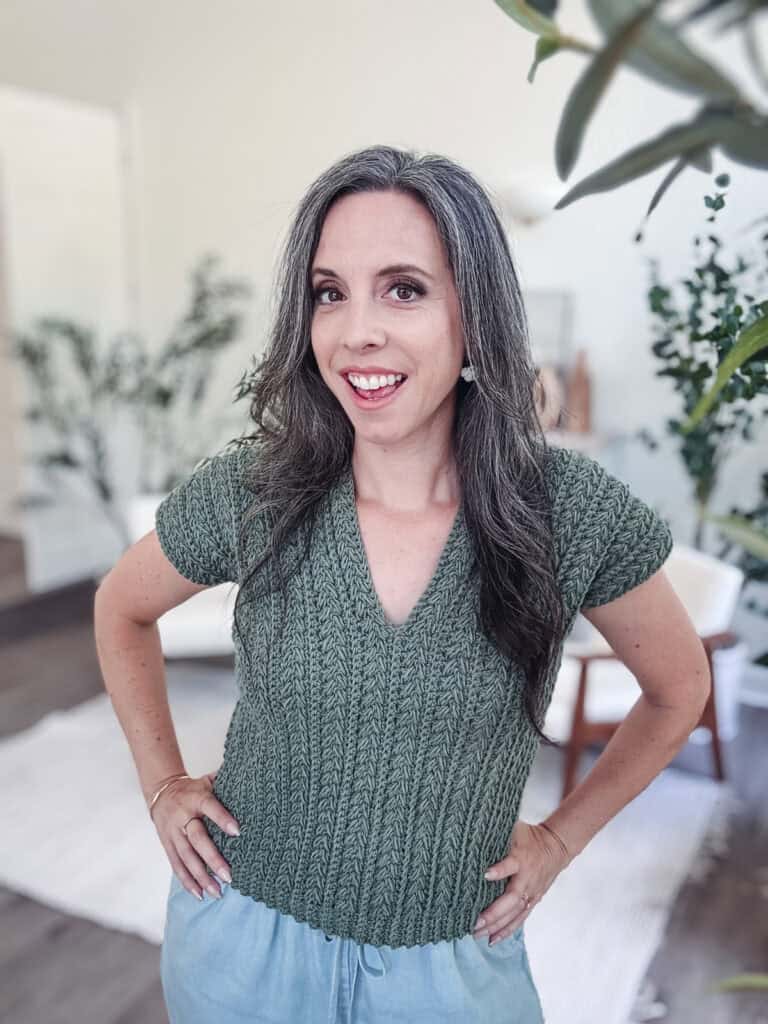 A woman with long, gray hair and a green Braided Wheat Easy Summer Crochet Top Pattern stands indoors, smiling at the camera with her hands on her hips. The background shows plants and light-colored furniture.