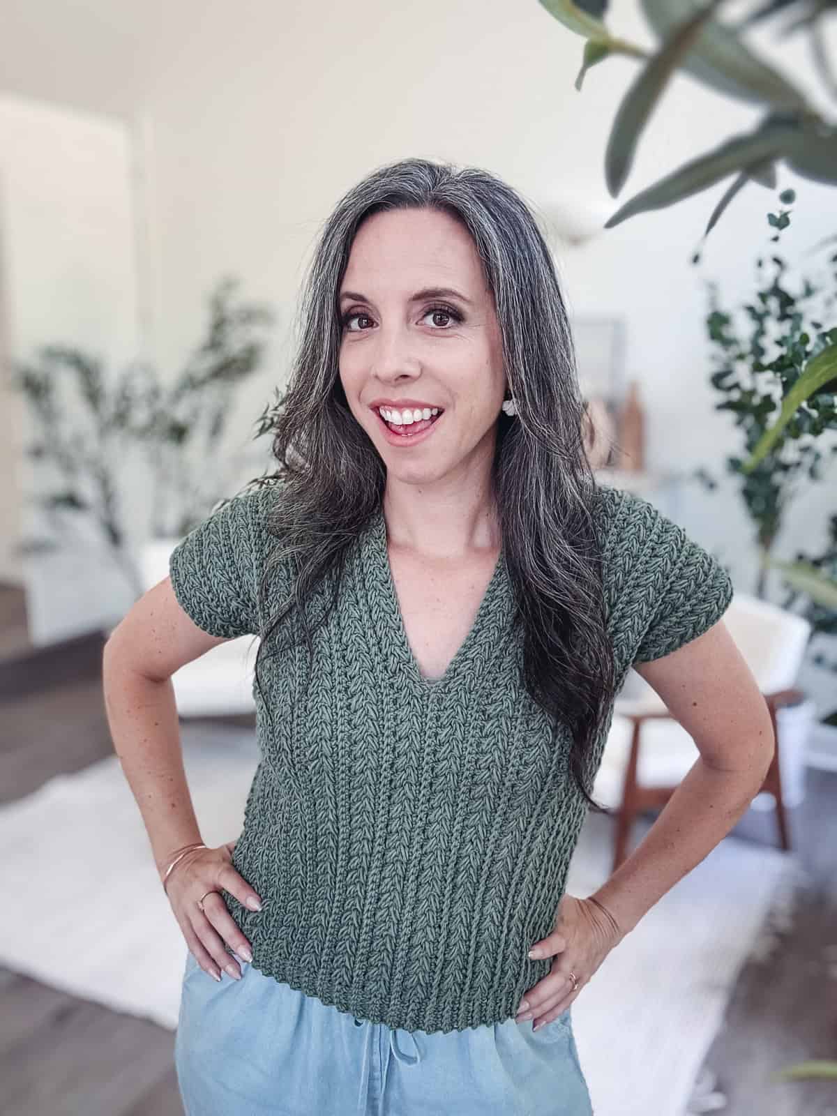 A woman with long, gray hair and a green Braided Wheat Easy Summer Crochet Top Pattern stands indoors, smiling at the camera with her hands on her hips. The background shows plants and light-colored furniture.