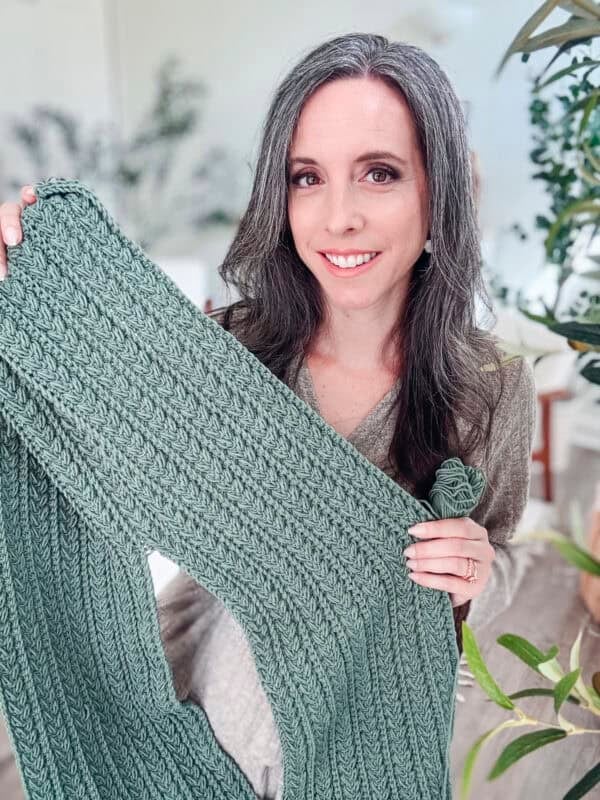A person with long dark hair holds up a knitted green scarf, smiling. They are in a bright room with plants.