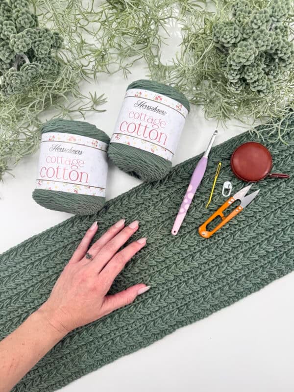 A hand rests on a green knitted fabric piece on a white surface, surrounded by green yarns and various crochet tools.