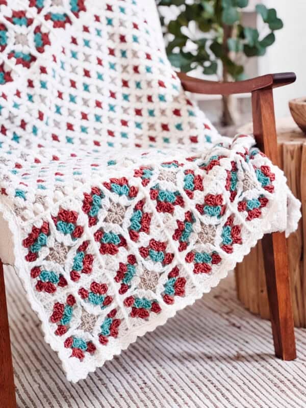 A crocheted afghan featuring the Free Crochet Pattern: Windowpane Granny Stitch Blanket, with a multicolored granny square pattern in white, red, teal, and beige, draped over a wooden chair.