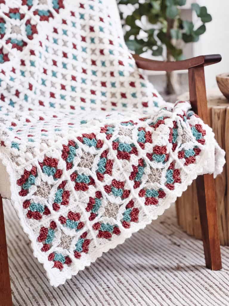 A crocheted afghan featuring the Free Crochet Pattern: Windowpane Granny Stitch Blanket, with a multicolored granny square pattern in white, red, teal, and beige, draped over a wooden chair.