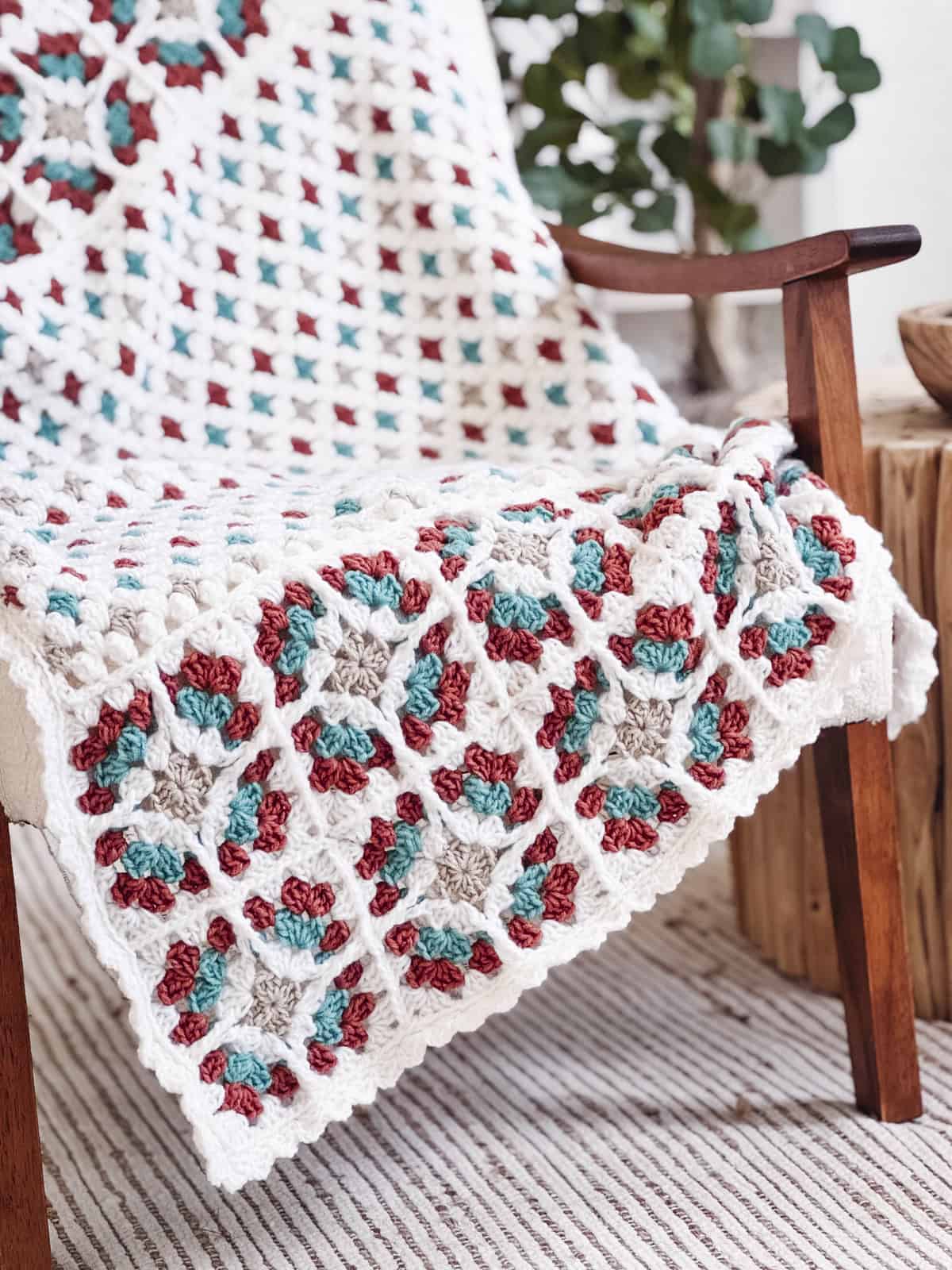 A crocheted afghan featuring the Free Crochet Pattern: Windowpane Granny Stitch Blanket, with a multicolored granny square pattern in white, red, teal, and beige, draped over a wooden chair.