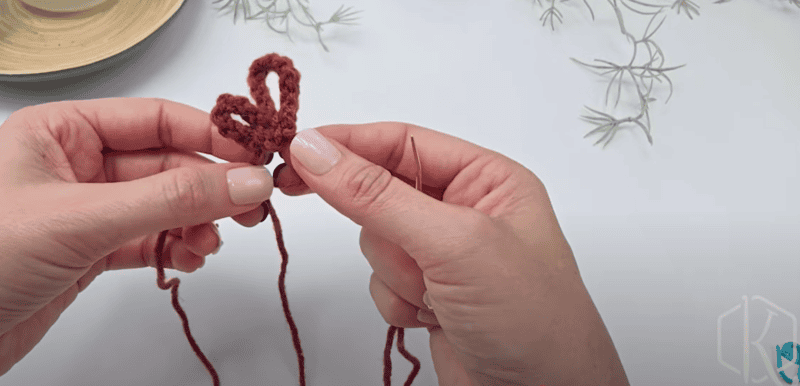 Two hands holding a purple crocheted heart shape with a yarn needle and loose yarn, on a white surface with green plant decorations.