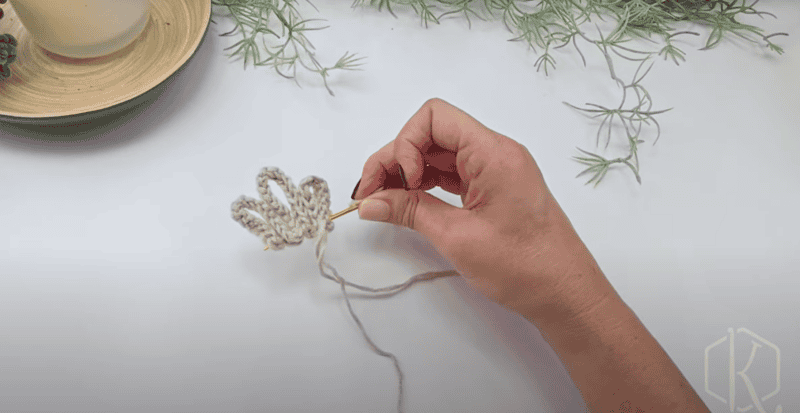 A hand holds a crochet hook and yarn, working on a scalloped crocheted piece, with greenery and a candle in the background on a white surface.