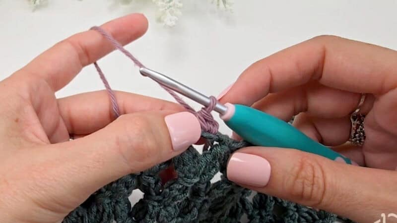 Close-up of hands using a teal-handled crochet hook to work with purple and green yarn, demonstrating how to crochet a clean, straight edge.
