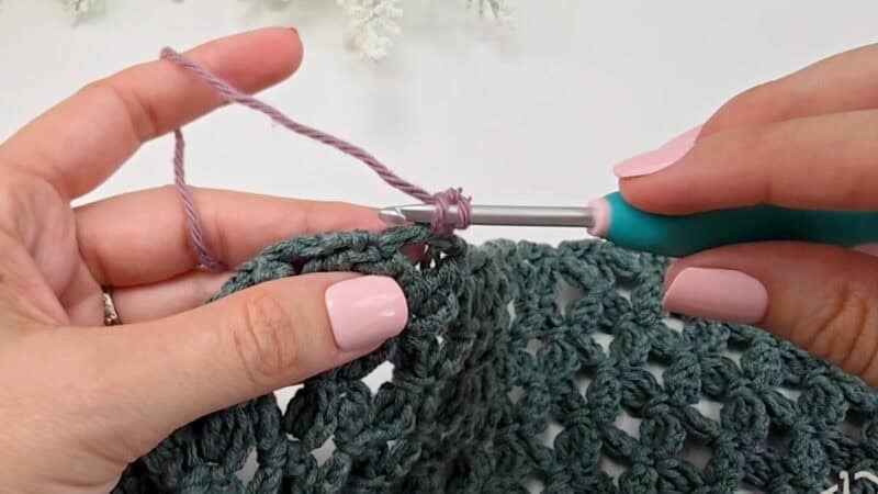 Close-up of hands crocheting with a green hook and purple yarn, working on a partially completed textured green crochet piece—perfect for those learning how to crochet a clean, straight edge.