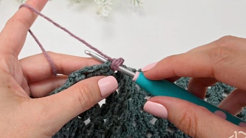 Close-up of hands crocheting with a teal crochet hook and purple yarn, working on a green textured fabric with a straight edge, against a white background.