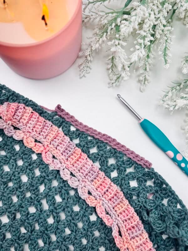 A crochet project with multicolored yarn, a crochet hook, and a lit pink candle are arranged on a white surface with frosted greenery, inspiring ideas on How to Crochet a Clean, Straight Edge.
