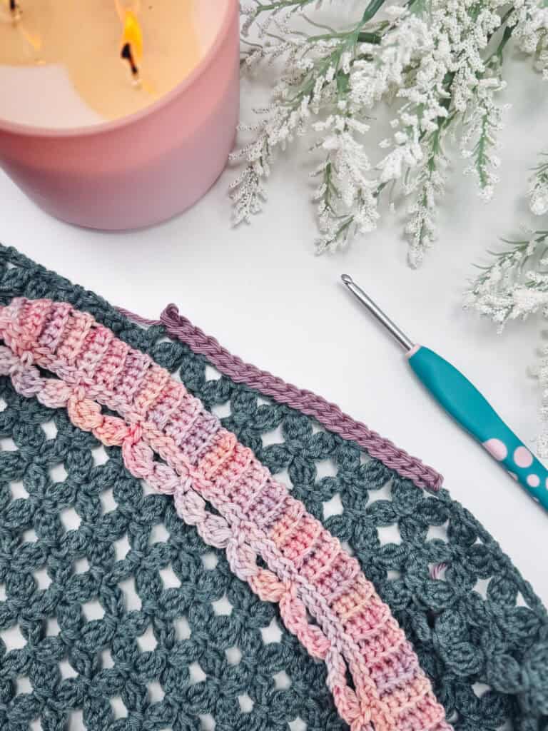 A crochet project with multicolored yarn, a crochet hook, and a lit pink candle are arranged on a white surface with frosted greenery, inspiring ideas on How to Crochet a Clean, Straight Edge.