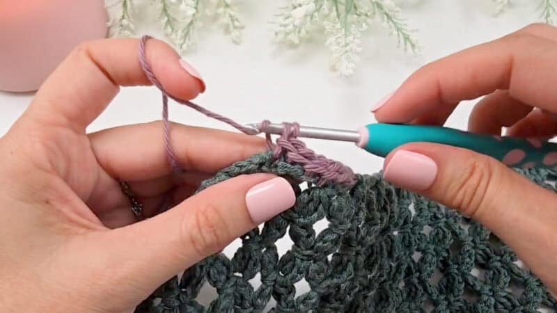 Close-up of hands demonstrating how to crochet a clean, straight edge with a teal and white crochet hook, working purple yarn on textured green fabric. White decorative foliage adorns the background.