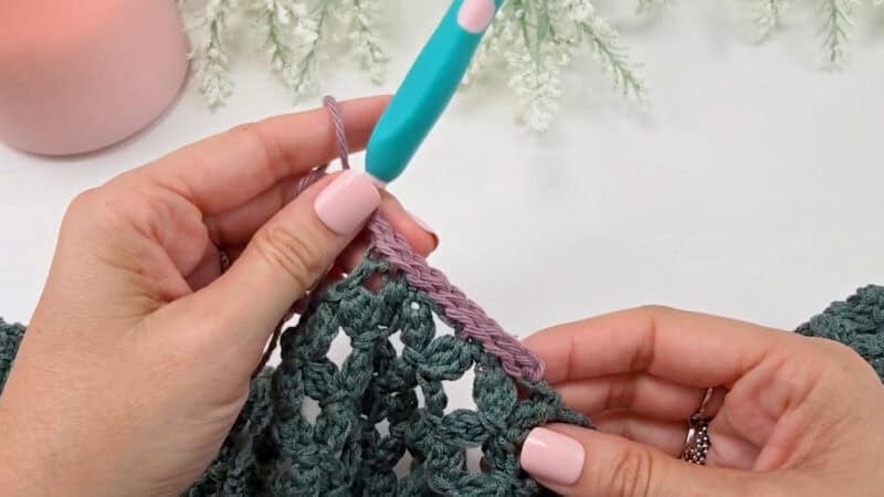 Hands crocheting with a teal crochet hook, adding a row of lavender yarn to a green textured crochet project, with decorative greenery in the background—perfect for those learning how to crochet a clean, straight edge.
