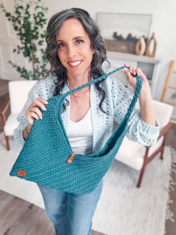 Woman with wavy gray hair in a light blue sweater holds up a teal Wheat Stitch Crochet Rectangle Bag in a bright, modern living room.