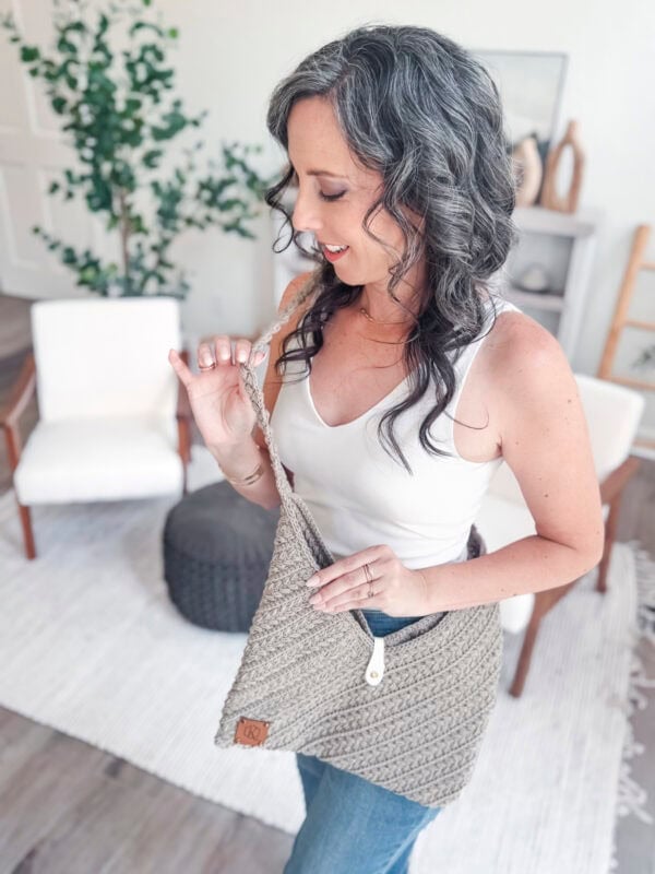 A woman with wavy gray hair stands indoors, wearing a white sleeveless top and blue jeans, holding a Wheat Stitch Crochet Rectangle Bag. Modern, minimalistic decor is visible in the background.