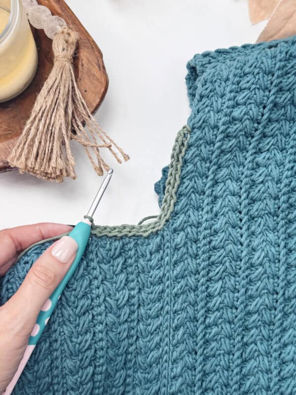 A hand holds a crochet hook, working with green yarn on the neckline of a textured blue garment. A tassel and candle are nearby, as the crafter demonstrates how to crochet a clean, straight edge for a polished finish.