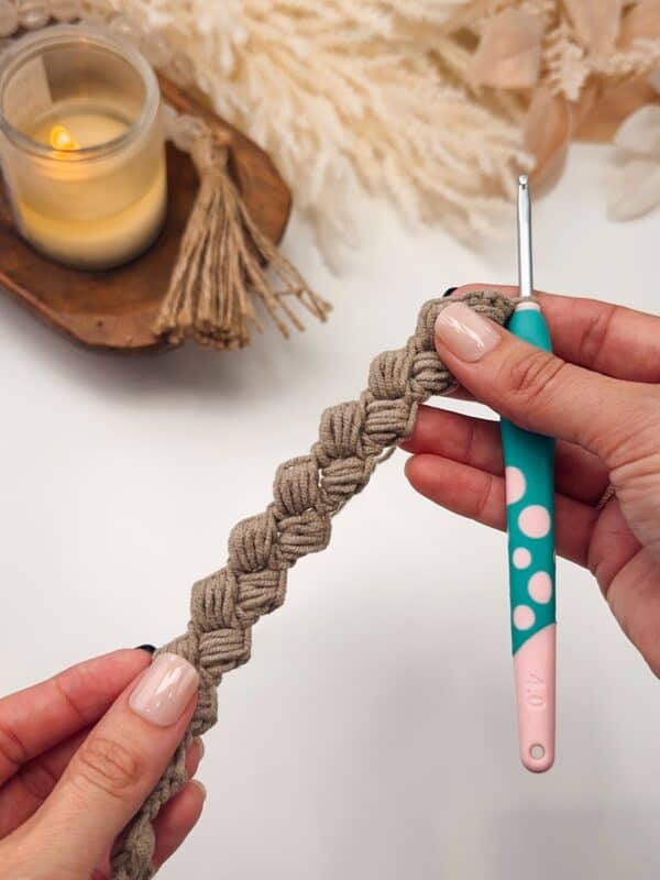 A person holds a crocheted beige band and a crochet hook, with a lit candle and decorative items in the background.