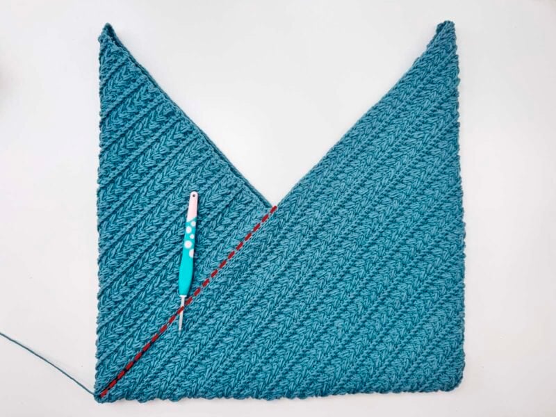 A textured blue crochet piece forms a V shape with a crochet hook placed on top; a red dotted line marks one side.