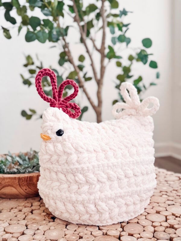 A soft, white, knitted chicken with a red comb and white tail sits on a wooden table, with a plant and a bowl in the background.