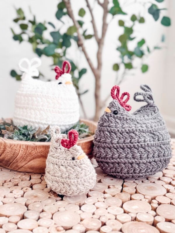 Three crocheted chickens in white, beige, and gray are displayed on a wooden surface with a small plant in the background. Make your own with the Free Wheat Stitch Chickens Crochet Pattern.
