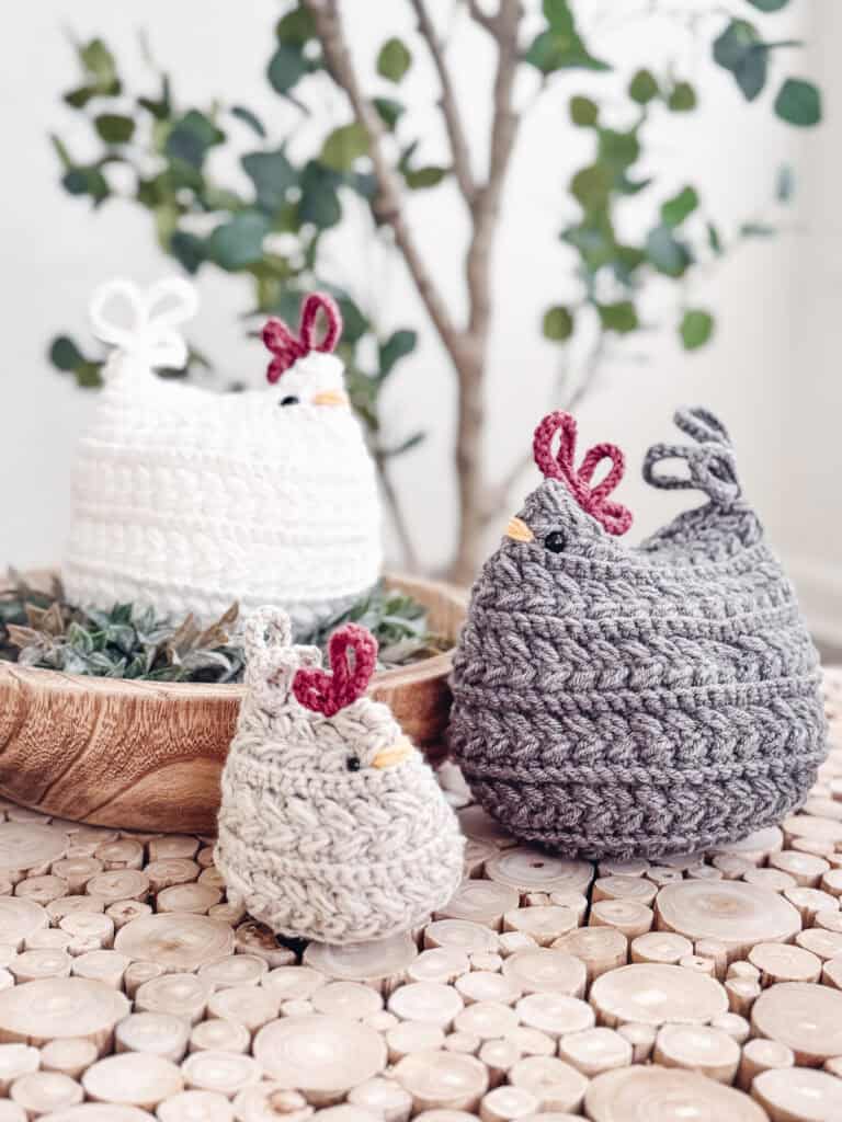 Three crocheted chickens in white, beige, and gray are displayed on a wooden surface with a small plant in the background. Make your own with the Free Wheat Stitch Chickens Crochet Pattern.