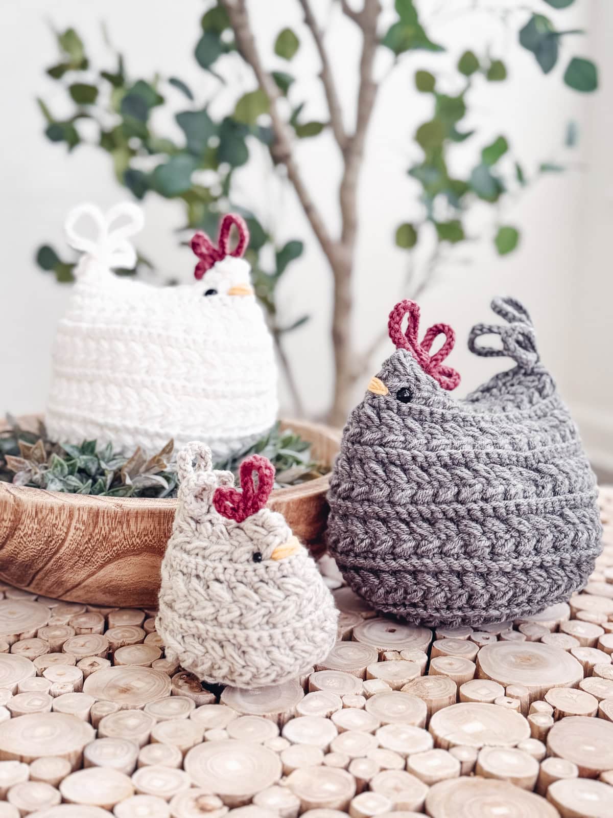 Three crocheted chickens in white, beige, and gray are displayed on a wooden surface with a small plant in the background. Make your own with the Free Wheat Stitch Chickens Crochet Pattern.