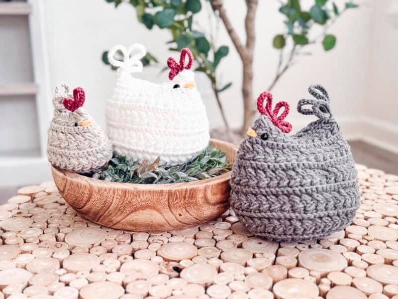 Three crocheted chicken figures in white, gray, and beige are displayed in and beside a wooden bowl on a round wooden table, with a green plant in the background.