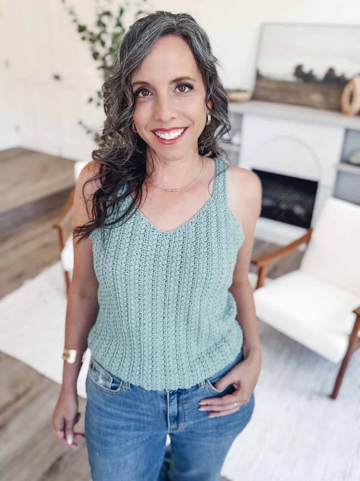 A woman with wavy gray hair wearing a light blue knitted tank top—crafted from modern crochet & knit patterns—and jeans stands indoors, smiling at the camera.