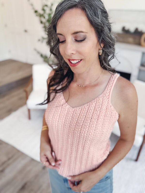 Size inclusive crochet tank top xs 5x Woman with wavy gray hair wearing a light pink knit tank top and blue jeans stands indoors, looking down with a slight smile. The background shows home furnishings and a plant.