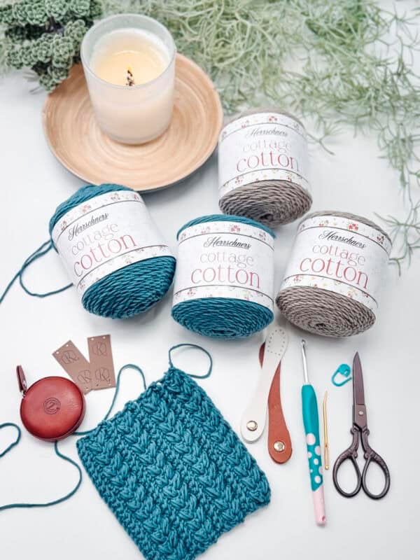 Four skeins of cottage cotton yarn, crochet tools, scissors, a candle, and a crocheted swatch are arranged on a white surface with greenery in the background.