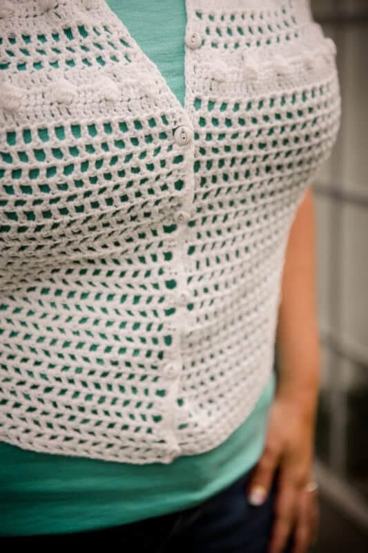 A person wearing a teal shirt and a white, crocheted vest with buttons, shown from the torso down.