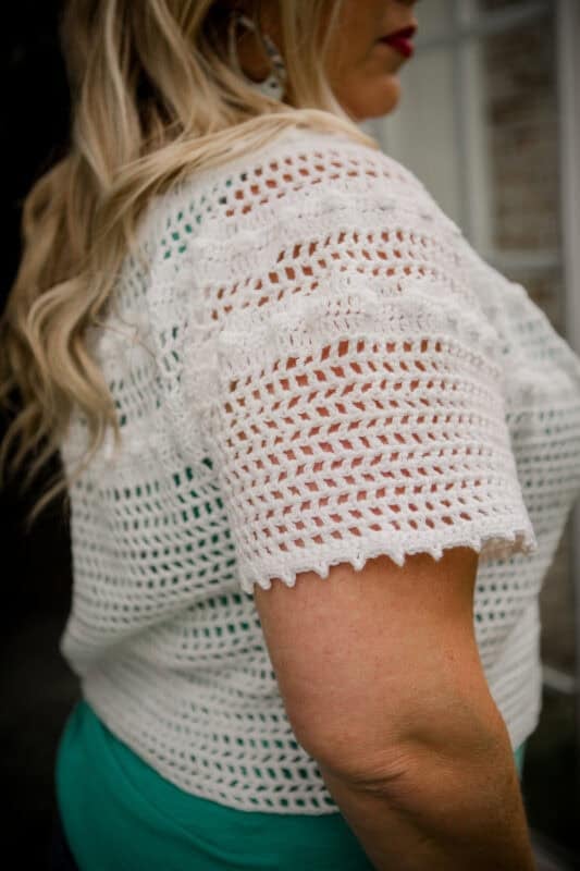 A woman with long blonde hair wearing a crocheted white short-sleeve top over a green shirt, shown from the side.