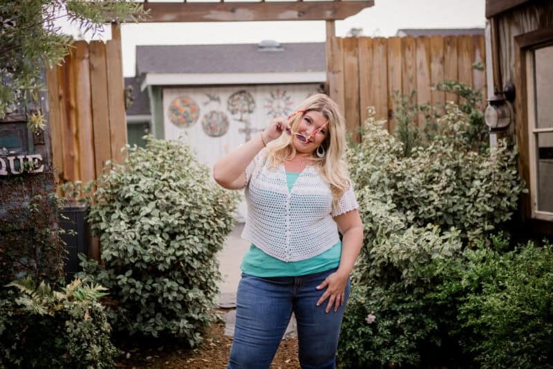 A woman stands outdoors in front of a wooden fence and greenery, smiling and making a peace sign with her right hand near her face.