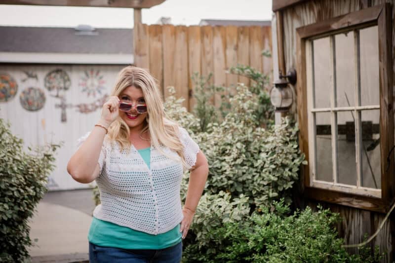 Woman with long blonde hair wearing sunglasses and a white crochet top stands outside near a wooden fence and window, looking at the camera.