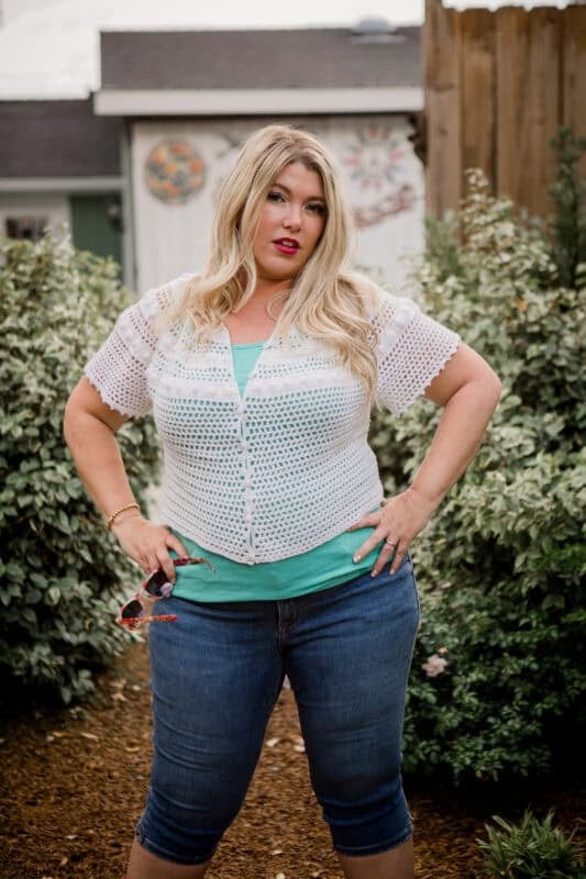 A woman with long blonde hair stands outdoors in front of greenery, wearing a white crochet top over a green shirt and blue denim shorts, holding sunglasses in one hand.