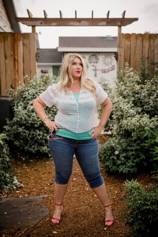 A woman with long blonde hair stands in a garden, wearing a white crochet top, teal tank, denim capris, and red heels, with her hands on her hips and looking to the side.